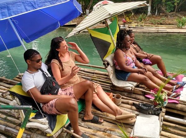 Four casually dressed travelers are seated on two bamboo rafts waiting for the driver to shuttle them down the White River area that spans the St. Ann and St. Mary parishes of Jamaica. Two of these guests have traveled here with Island Turf Tours and all are eagerly anticipating the ride. Jamaica flags grace both rafts, along with small bits of floral decorations to make it appear as though those who are seated are floating along on a wooden garden.